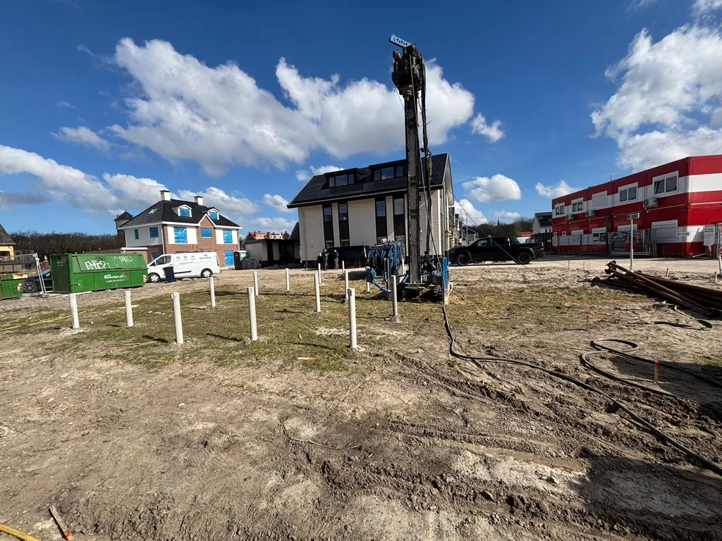 Bouwkavel met een rij uitstekende witte buizen en een boorstelling voor casing draaipalen op de achtergrond