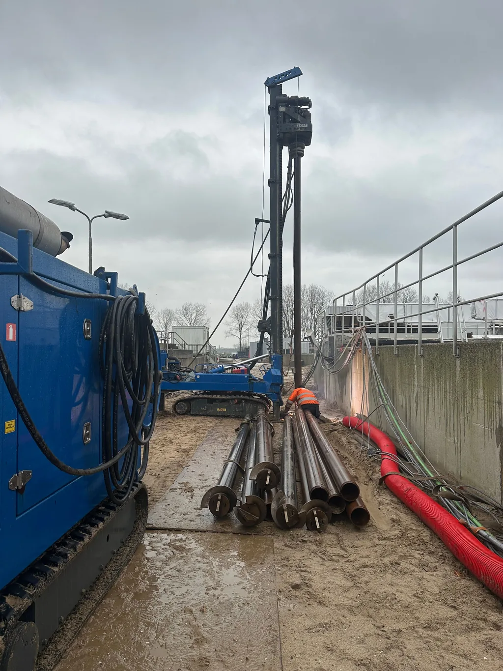 Stalen buizen voor casing draaipalen liggen klaar naast twee boorstellingen langs een betonnen wand