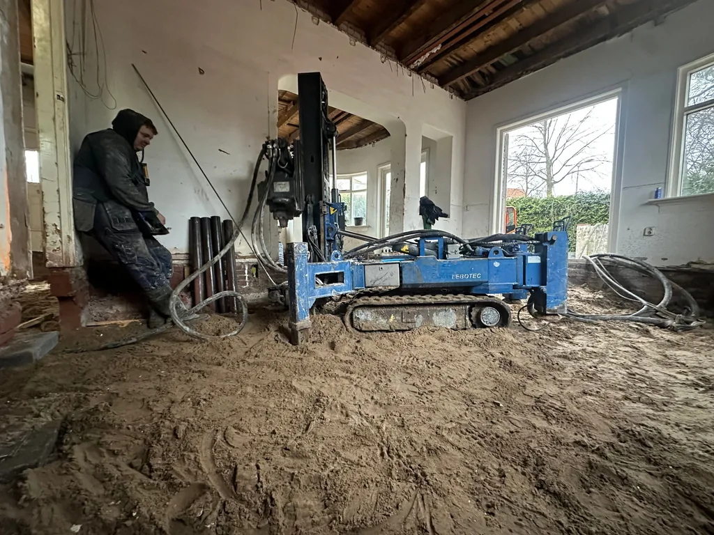 Compacte boormachine werkt inpandig aan funderingsherstel in een gestripte woonkamer met zandvloer en stalen buizen