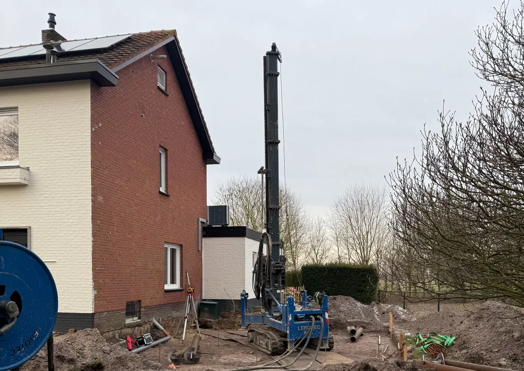 Blauwe boorstelling voor casing draaipalen naast een bakstenen woning in Essen, België, met uitgegraven grond voor de aanbouw