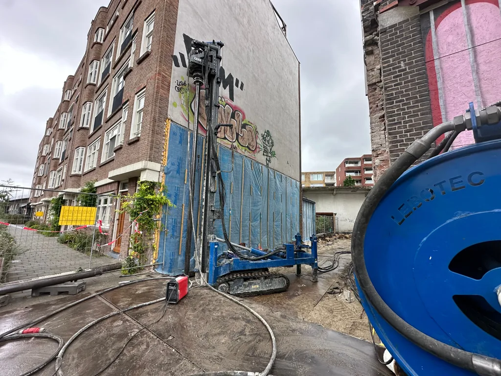 Boorstelling voor casing draaipalen tegen een afgeschermde blinde gevel in Rotterdam, met slanghaspel op de voorgrond