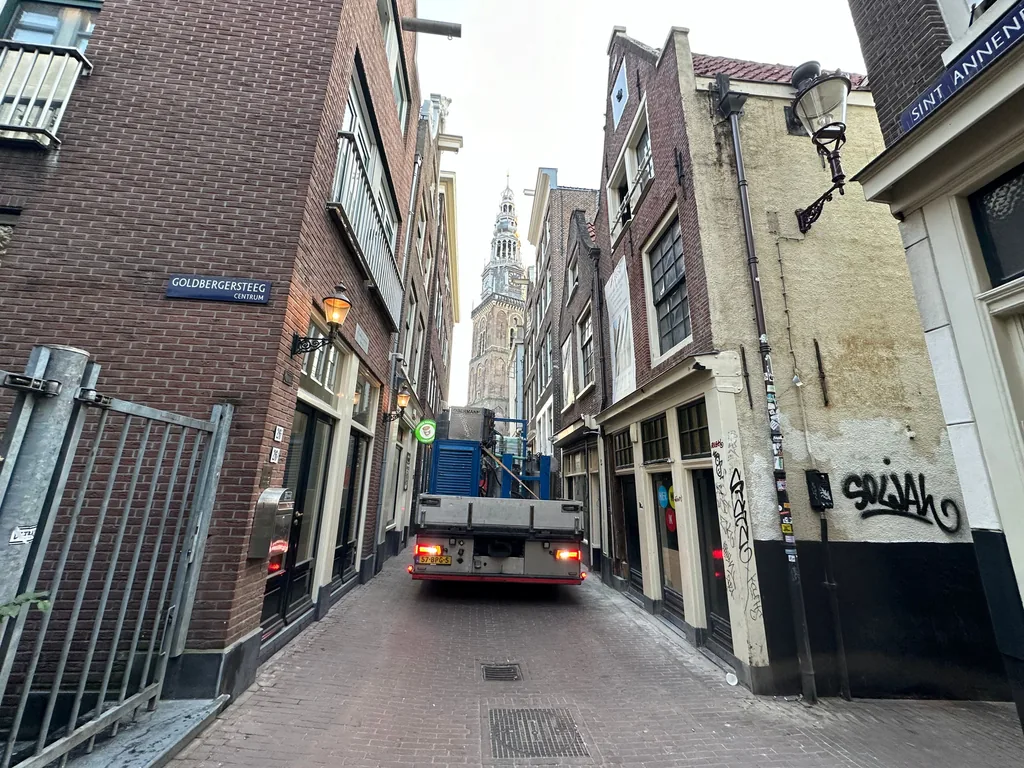 Vrachtwagen voert materieel voor schroefinjectiepalen aan door de smalle Goldbergersteeg bij de Sint Annendwarsstraat in Amsterdam, met de Oude Kerk op de achtergrond