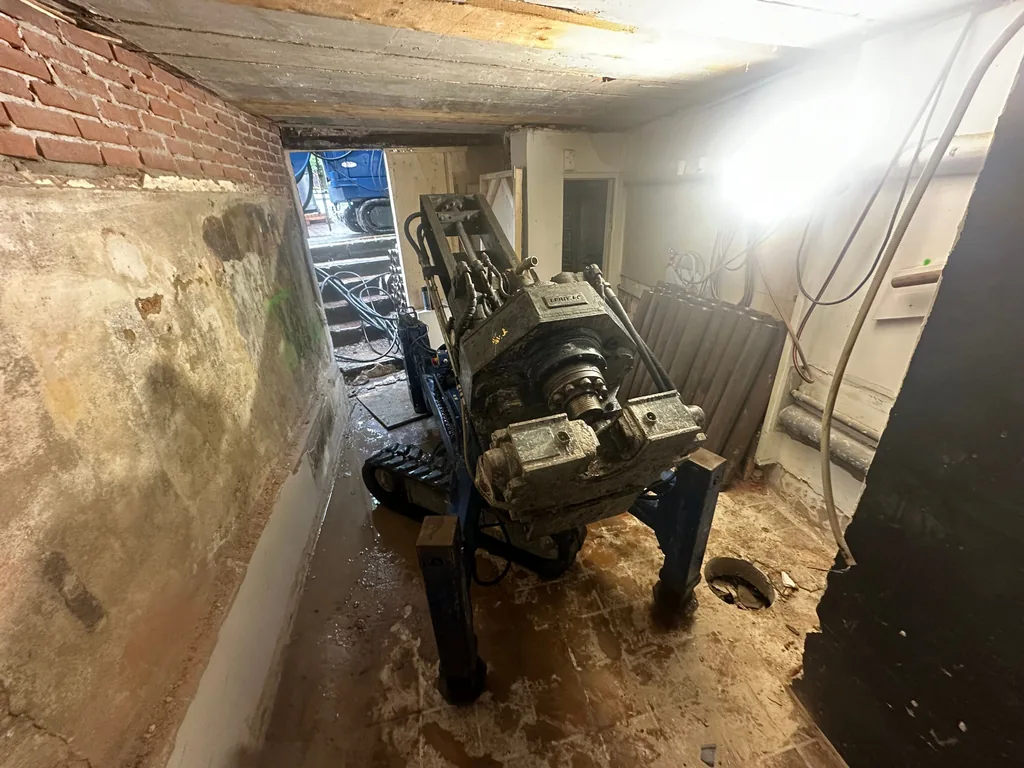 Compacte boorstelling voor schroefinjectiepalen in een smalle gang van een pand in hartje Amsterdam, met de toegangstrap op de achtergrond