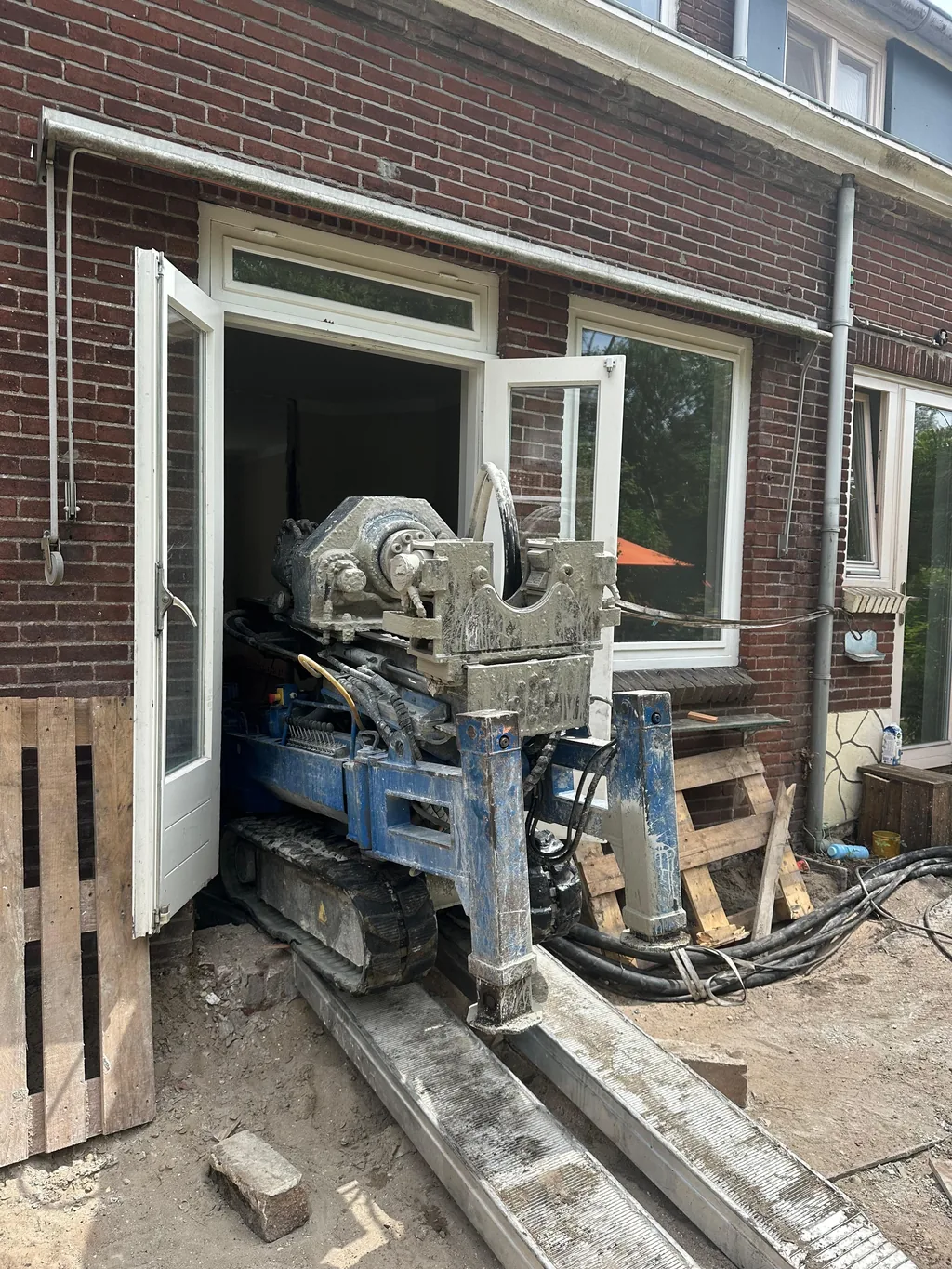 Compacte boorstelling rijdt via loopplanken door de tuindeuren een rijtjeshuis in Duivendrecht binnen voor schroefinjectiepalen