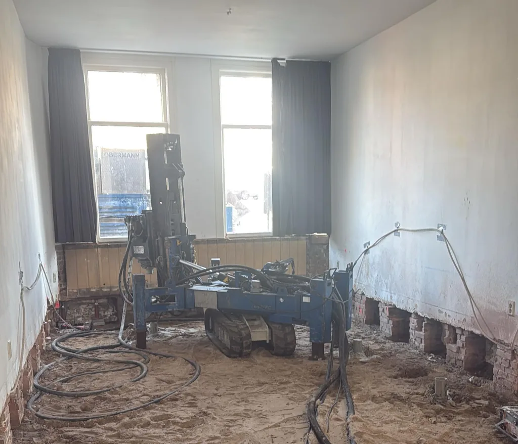 Boorstelling boort schroefinjectiepalen in een gestripte kamer aan de Kampersingel in Haarlem, met vrijgegraven muurvoeten langs de wand