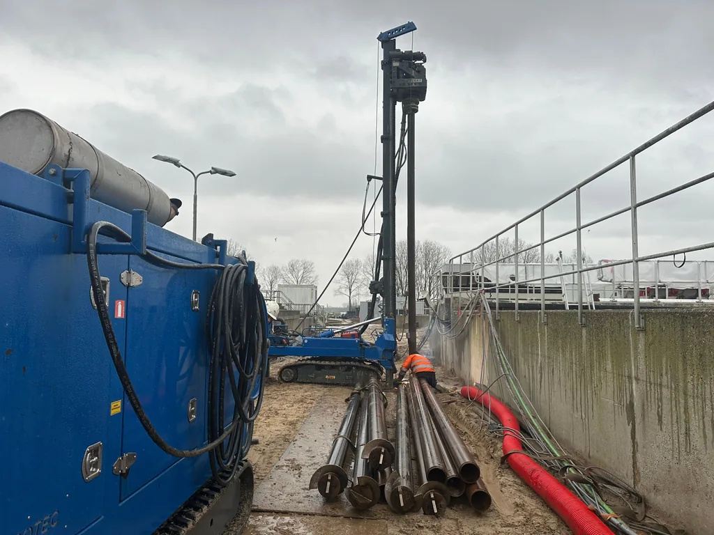 Boorstelling en stapel paaldelen voor schroefinjectiepalen langs een betonnen bak op de RWZI in Oijen