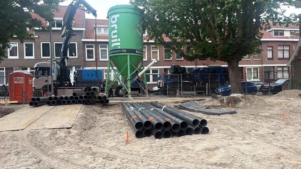 Stapels stalen buizen voor schroefinjectiepalen, een kraanwagen en een groene mortelsilo op de bouwplaats in Rotterdam