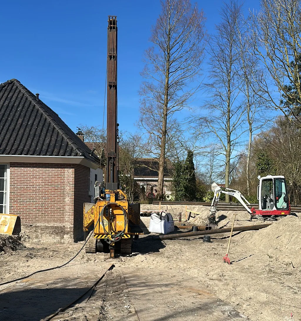 Heistelling voor stalen buispalen naast een woning in Heerhugowaard, met minigraver en buizen op het terrein