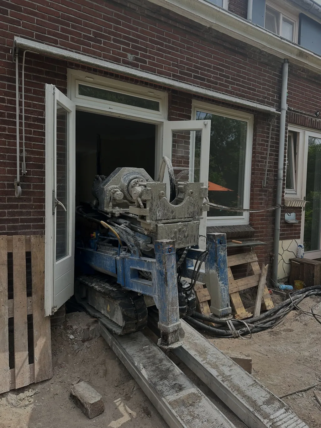 Compacte boorstelling voor schroefinjectiepalen rijdt via planken door openslaande tuindeuren een woning binnen