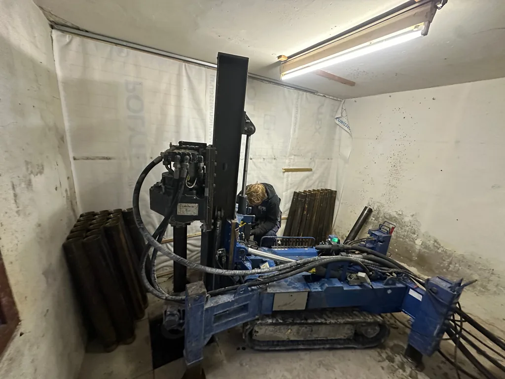 Medewerker bedient een compacte boorstelling in een kleine afgeschermde ruimte met stapels buisdelen voor schroefinjectiepalen