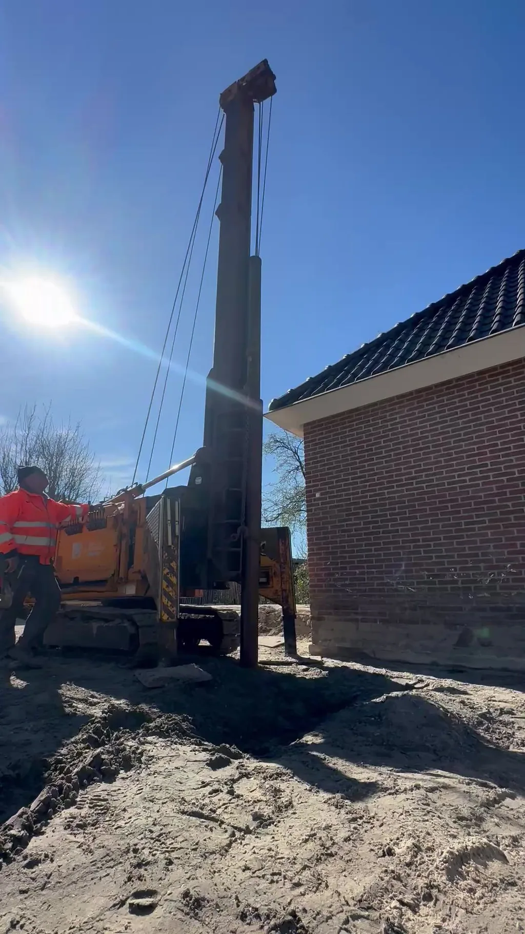 Heistelling op rupsen heit een stalen buispaal in het zand vlak naast de bakstenen gevel van een woning, met een medewerker in oranje werkkleding ernaast